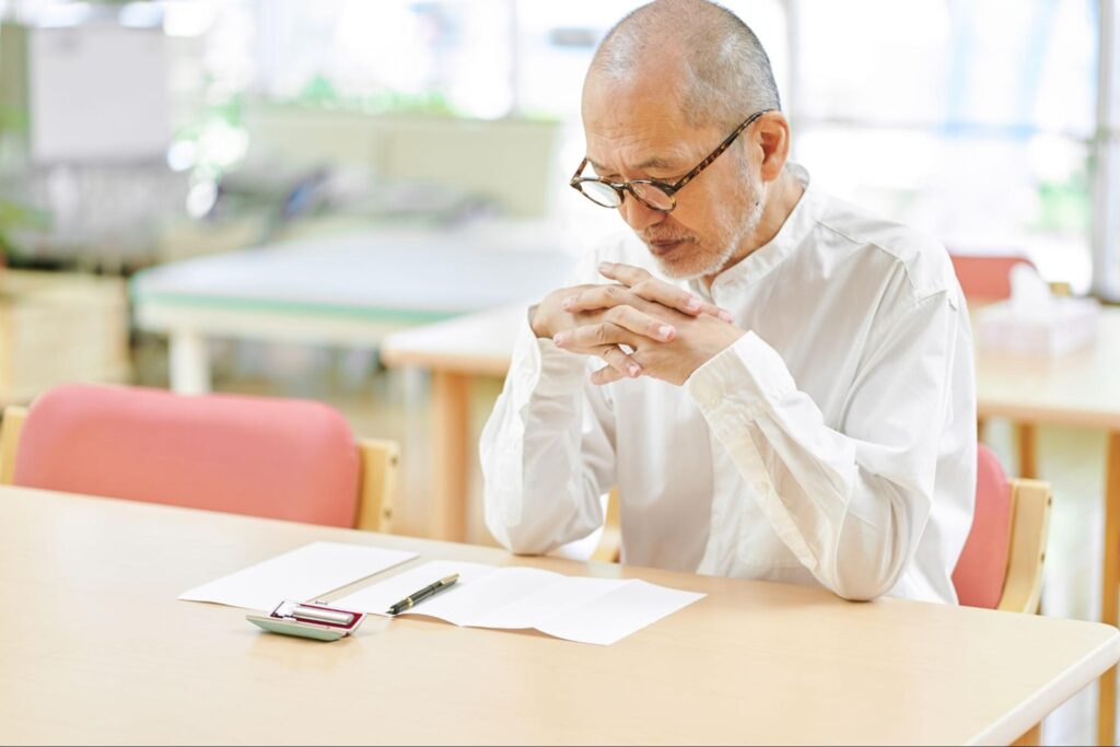 誤嚥性肺炎が再発してしまったのか、テーブルに座って深刻な顔をする高齢男性の画像