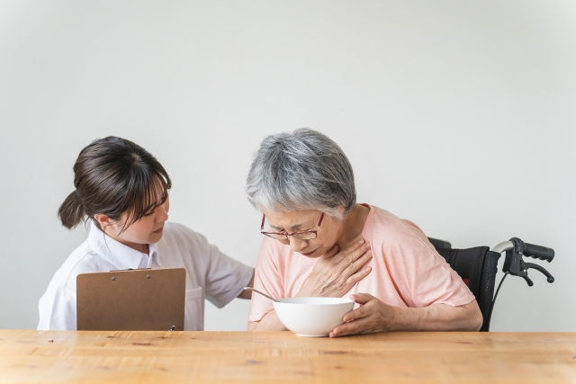 どんぶりの前で胸を押さえて苦しそうな高齢女性と背中をさする医療従事者の女性