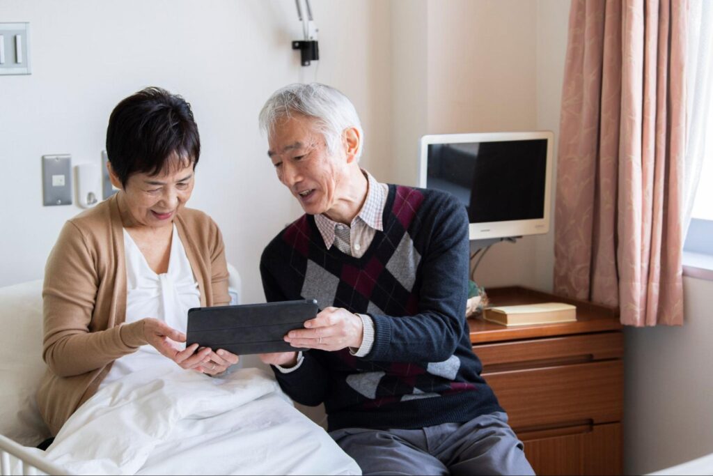 入院中の高齢女性にタブレットらしきものを見せる高齢男性